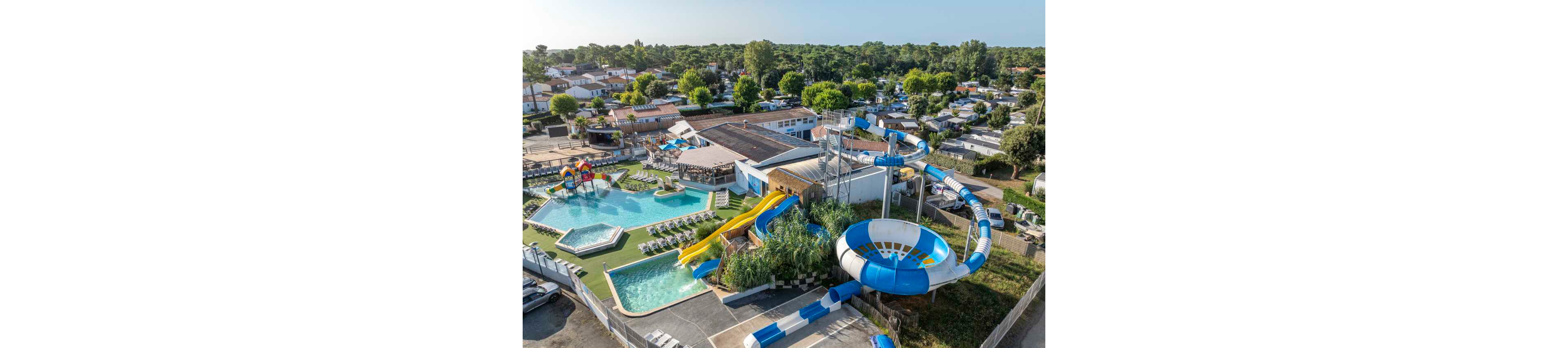A large outdoor water park is shown from above within a busy campsite or holiday resort. Multiple swimming pools, including a main pool and a shallow children's pool with small slides, are surrounded by sun loungers and umbrellas. A tall structure supports twisting blue and white water slides and a large bowl slide feature. Nearby, rows of mobile homes and caravans are nestled among trees, creating a relaxed vacation atmosphere.