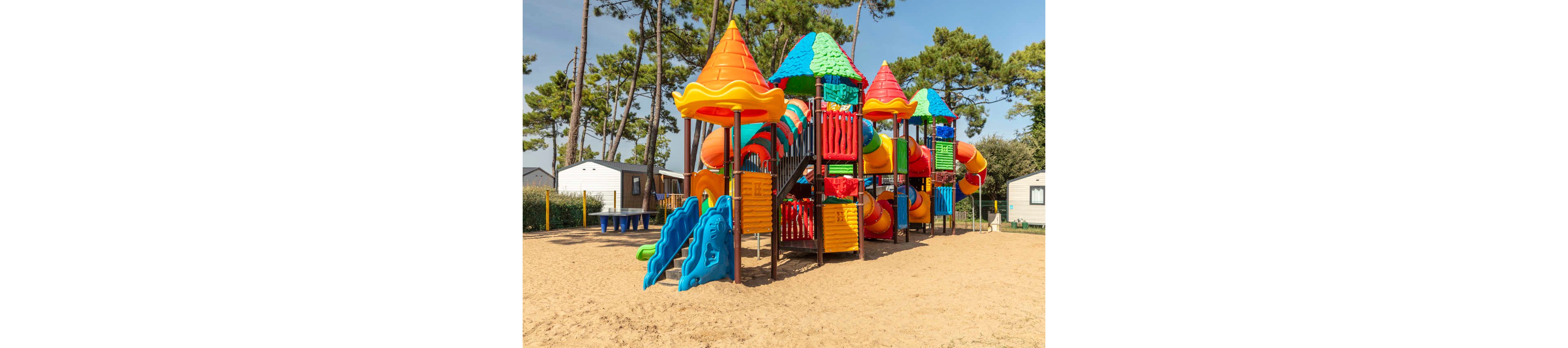 A large, colorful playground structure stands on a sandy surface in an outdoor park. The play area features multiple slides, tunnels, and towers with bright red, orange, yellow, blue, and green elements. Tall pine trees surround the playground, and small buildings or cabins are visible in the background. The scene is sunlit, suggesting a warm, pleasant day suitable for children’s outdoor activities.