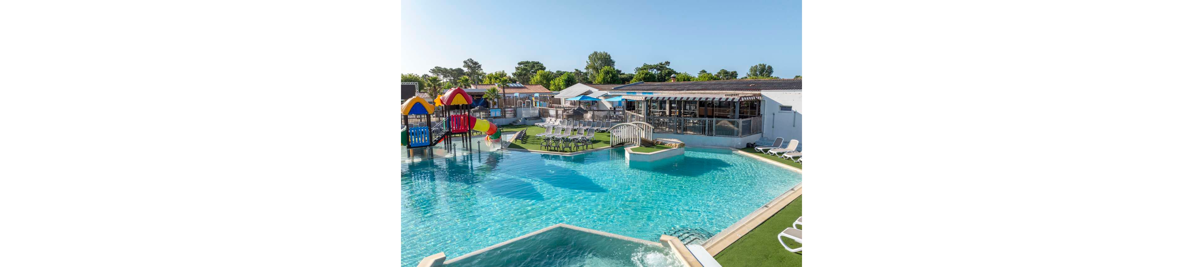 A large outdoor swimming pool is featured in a sunny resort setting. Colorful waterslides and a small play structure stand in the shallow area, suggesting a family-friendly environment. A small bridge crosses part of the pool, leading to a grassy island with neatly arranged lounge chairs. Additional sun loungers line the poolside, and low buildings and trees frame the background under a clear blue sky.