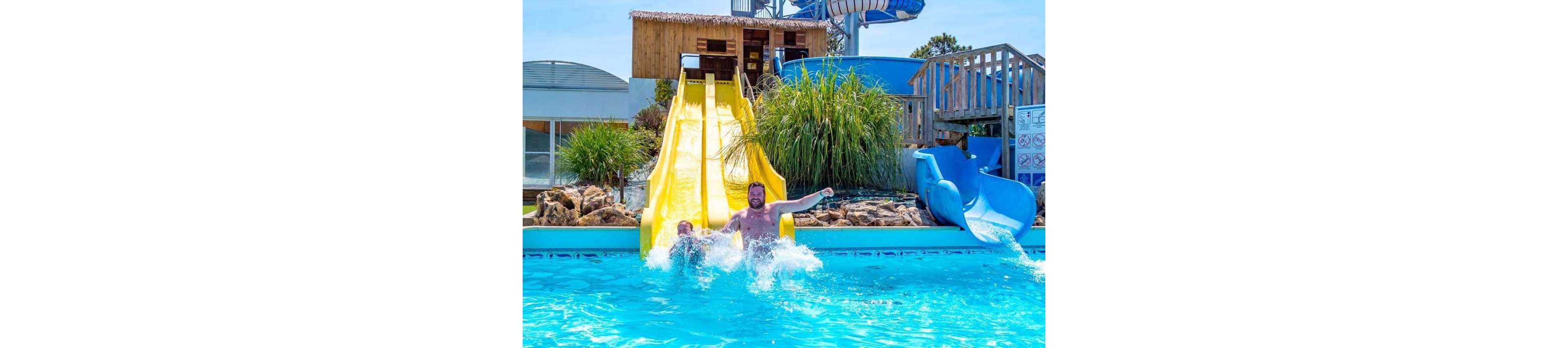 A man and child splash into a bright blue pool after riding a yellow water slide. The scene is set in an outdoor water park with additional slides and structures in the background. Green plants and rocks frame the slide area, adding a tropical feel. The sunny weather and clear sky create a fun, energetic atmosphere.