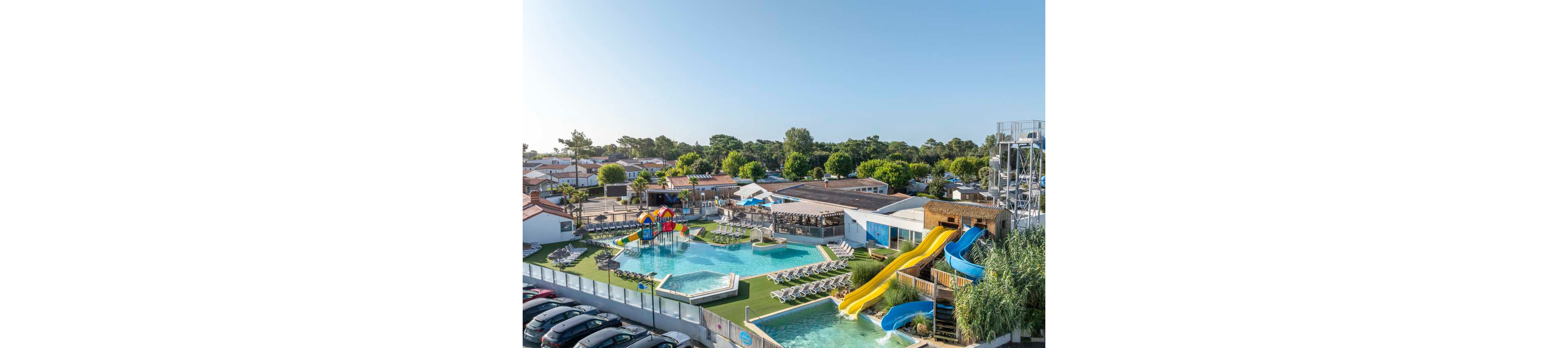 An aerial view shows a large family resort featuring multiple outdoor swimming pools and colorful waterslides. Sun loungers are neatly arranged around the pools, with a dedicated children's play area including small slides and water features. Several low-rise buildings and a bar or snack area surround the pool complex, while a parking lot with numerous cars sits in the foreground. Lush green trees and a clear blue sky create a bright, summery atmosphere.