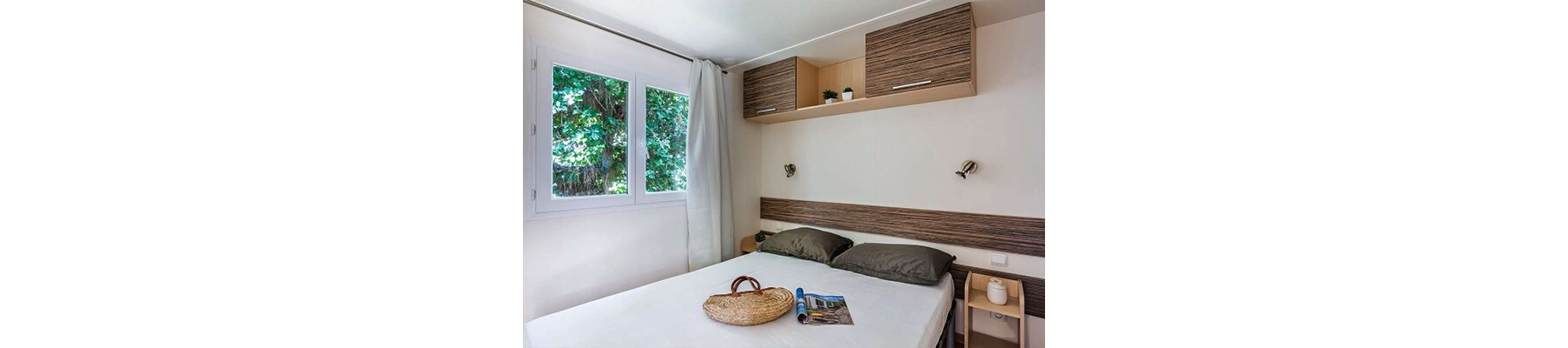 A small modern bedroom features a neatly made double bed with neutral bedding. A large window with white curtains lets in natural light and overlooks dense green foliage outside. Wooden cabinets and a headboard add warmth to the minimalist decor, with a small plant and decor items on the shelves. A woven bag and an open magazine rest on the bed, suggesting a relaxed vacation or weekend retreat.