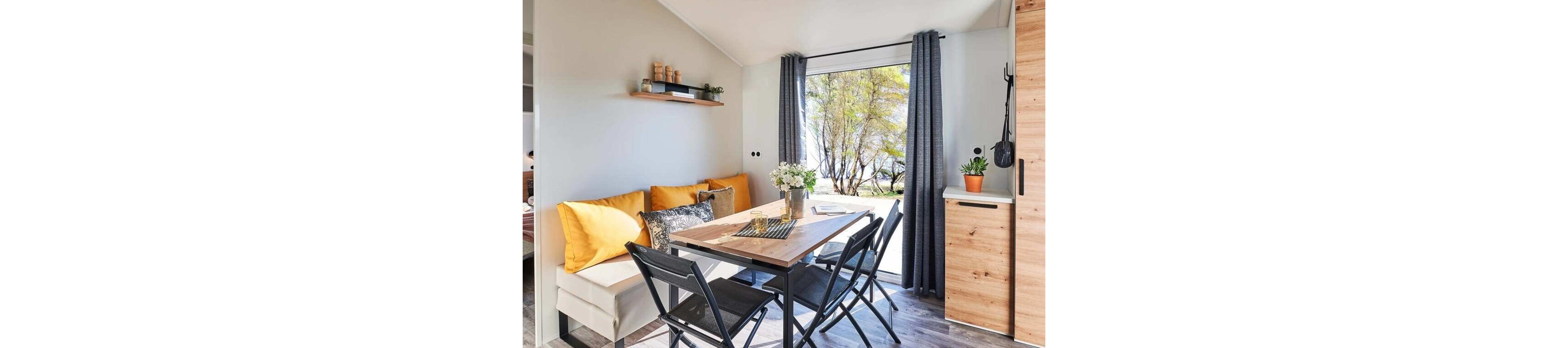 A cozy dining area features a wooden table with black folding chairs and a built-in bench with yellow cushions. Large glass doors with dark curtains open to a scenic outdoor view of trees and sunlight. A small vase of flowers decorates the table, while potted plants and jars on a wall shelf add natural accents. Light walls and wood finishes create a modern, airy atmosphere in the compact space.
