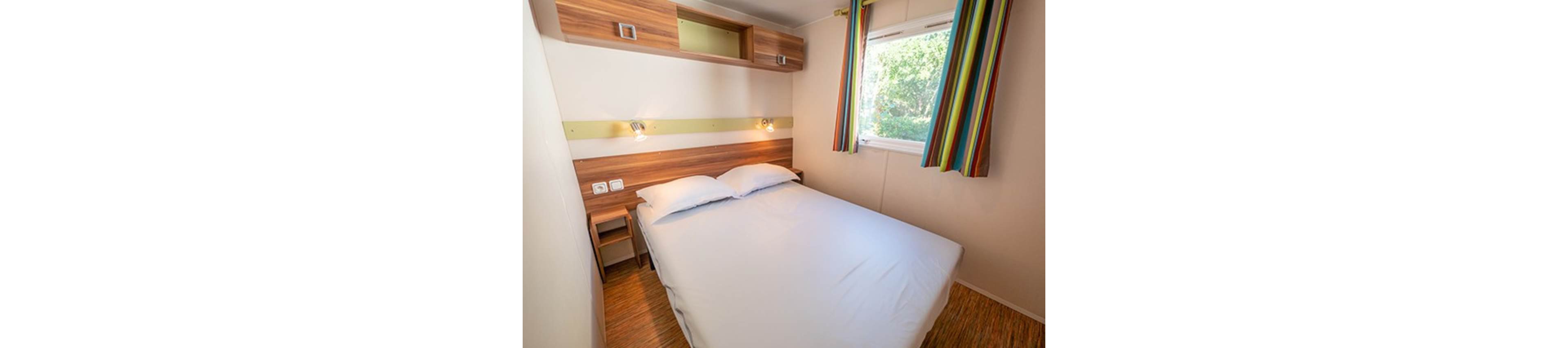Compact bedroom interior featuring a double bed with white linens and wooden headboard. Two wall-mounted reading lights illuminate the bed area, complemented by built-in wooden shelves and storage. A window with colorful striped curtains lets in natural light and offers a view of greenery outside. The room has a warm, minimalist design with light walls and wood-look flooring.