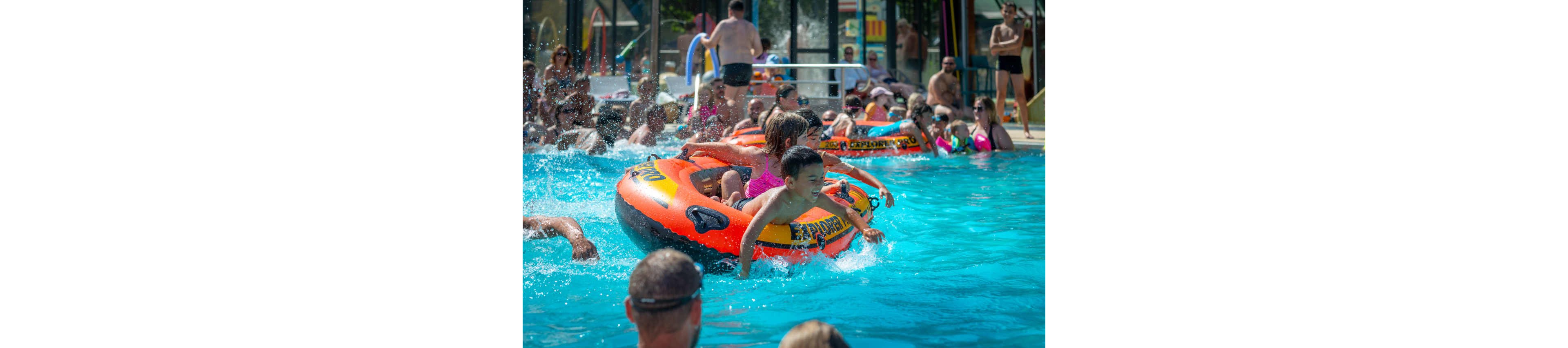 Children ride an orange inflatable raft in a crowded outdoor swimming pool. Adults and other kids relax and play in the bright blue water around them. Sunlight reflects off the splashing water, creating a lively summer atmosphere. The scene suggests a busy water park or public pool on a warm day.