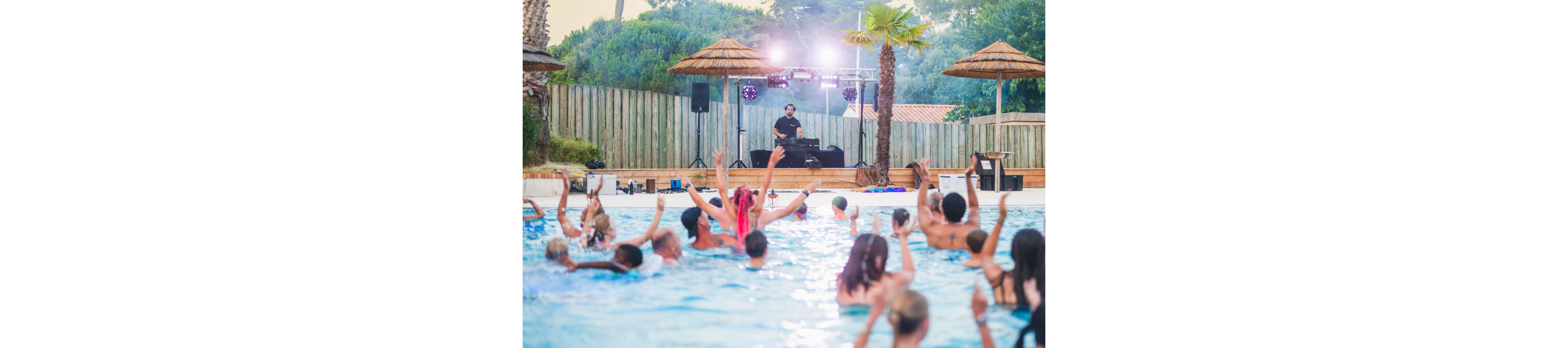 A DJ performs on a stage beside an outdoor pool while a crowd of people enjoy the water. Guests in the pool raise their hands and dance to the music, creating a lively party atmosphere. Palm trees, wooden fencing, and stage lights frame the scene, suggesting a resort or holiday setting. The overall mood is energetic, summery, and festive.