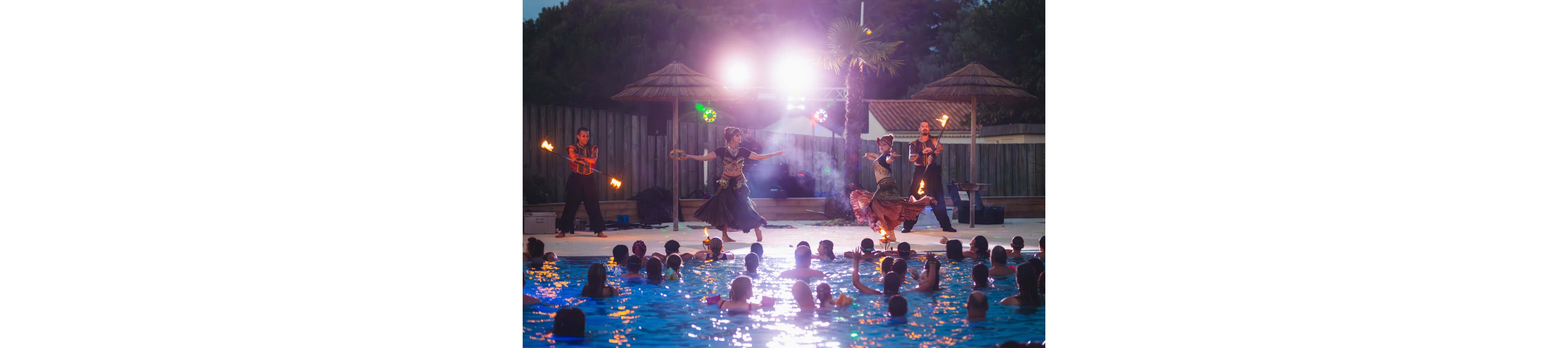 A group of fire dancers performs on a poolside stage at night. Swimmers and spectators watch from the illuminated swimming pool in the foreground. Bright stage lights, tiki-style umbrellas, and a wooden fence create a festive outdoor atmosphere. The scene captures motion, reflections on the water, and the dramatic glow of the flames.