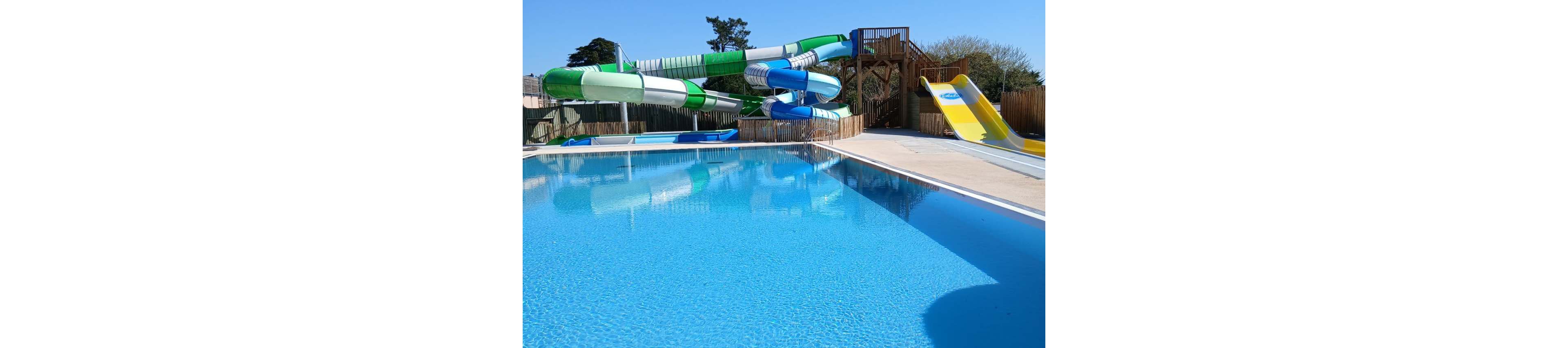 An outdoor swimming pool is shown with clear blue water reflecting the sky. In the background, several large twisting water slides in green, white, and blue curve down from a wooden platform. A wide yellow slide sits beside the main slides, leading into a shallow run-out area. The pool area is surrounded by fencing and trees, suggesting a leisure park or resort setting. Bright sunlight enhances the vibrant colors and creates a cheerful, summery atmosphere.