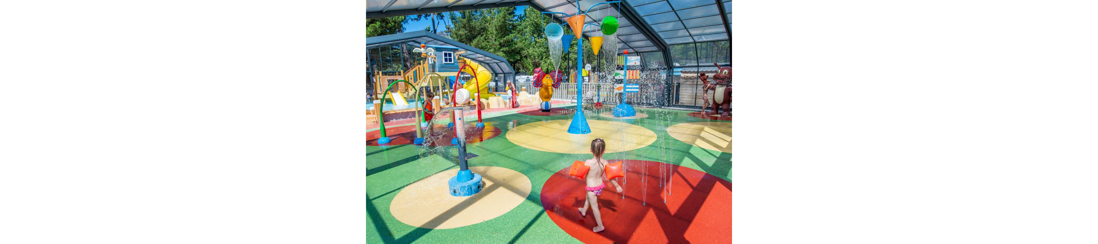 A young child wearing inflatable armbands runs through an indoor splash park. The play area features colorful circular patterns on the ground and multiple water fountains spraying from the floor. Overhead, large buckets and water features pour streams of water into the play zone. In the background, other children and adults enjoy slides, climbing structures, and additional water attractions under a transparent roof. Bright colors and playful designs create a fun, family-friendly atmosphere.