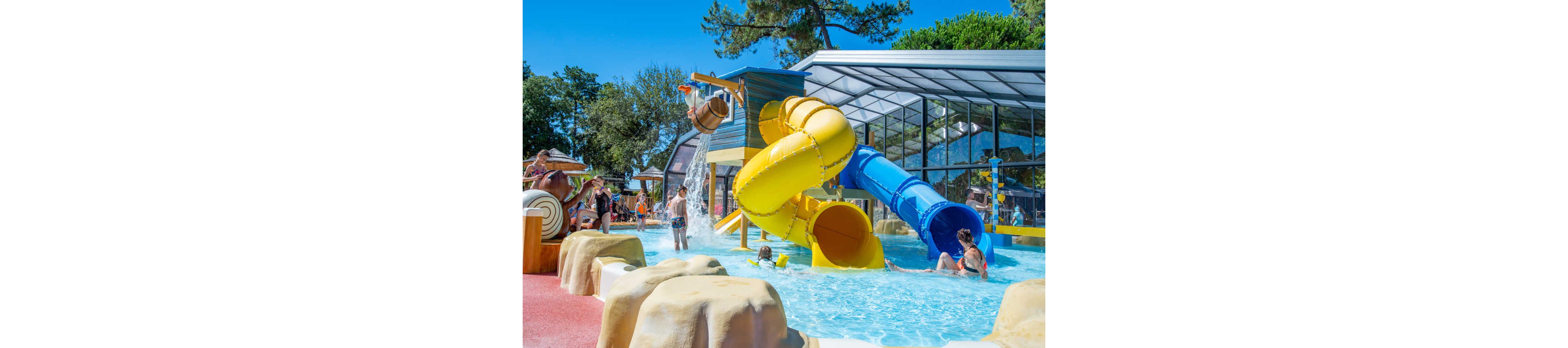 Children play in a shallow pool at a vibrant outdoor waterpark. A large yellow and blue spiral waterslide dominates the scene, ending in the clear blue water. A tipping bucket pours water onto kids below while others wait their turn near the structure. Adults and children relax around the pool area, with trees and a partially covered section in the background.