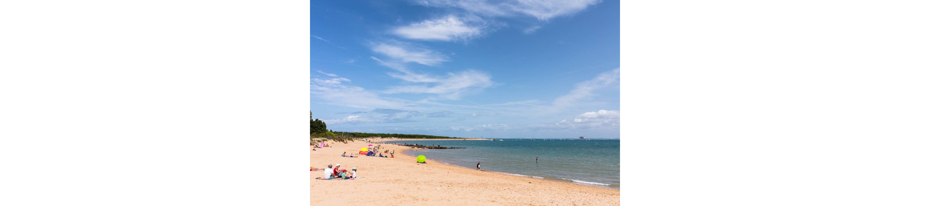 A wide sandy beach stretches along a calm blue sea under a bright, partly cloudy sky. Small groups of people relax on the sand with towels and umbrellas, while a few individuals wade in the shallow water. The shoreline curves gently toward a distant treeline and rocky outcrop. The scene conveys a peaceful summer day at a seaside destination.