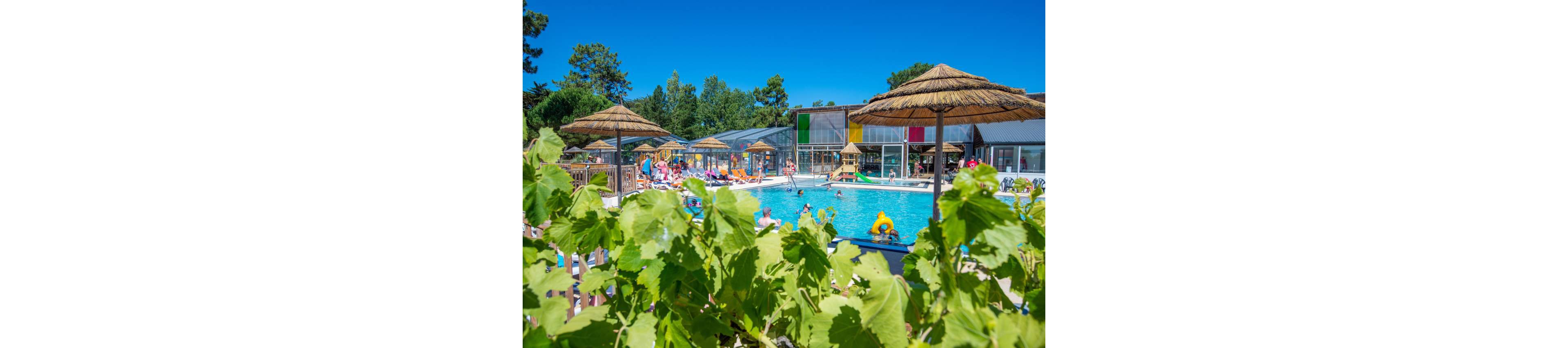 A lively outdoor swimming pool is surrounded by people relaxing and playing in the water. Straw parasols and wooden decking create a resort-like atmosphere under a clear blue sky. Green foliage in the foreground frames the scene, adding a fresh, natural touch. Colorful panels on the building and bright pool inflatables contribute to a cheerful summer holiday mood.