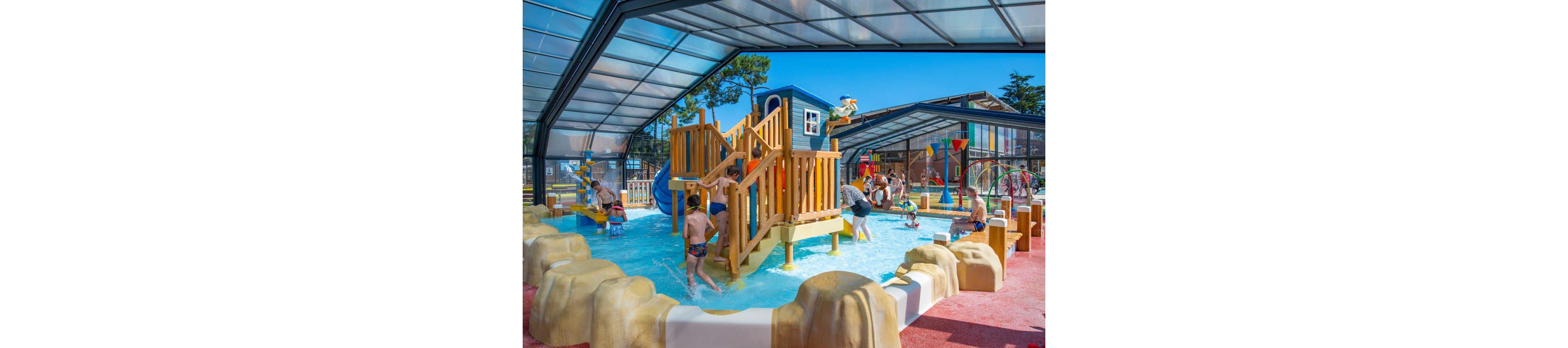 A group of children play on a colorful wooden and plastic climbing structure in a shallow indoor water playground. The pool area is covered by a large transparent roof, allowing bright daylight to fill the space. Kids climb stairs, slide down water slides, and splash in the shallow water while adults supervise nearby. The scene features playful decorations, including rocks, a small house-like structure, and water sprayers, creating a fun family-friendly atmosphere.