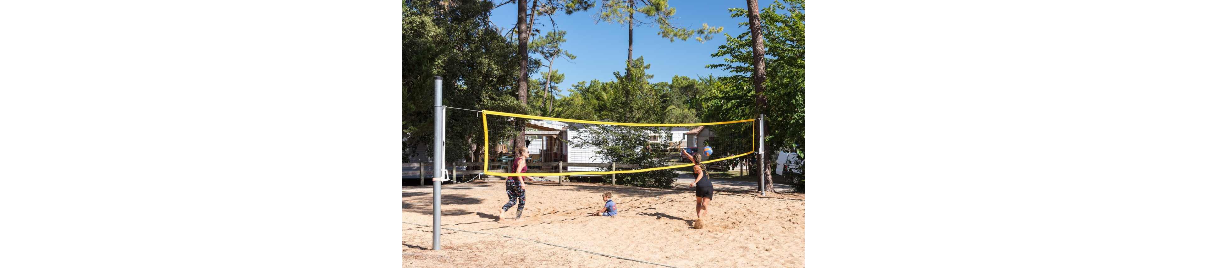 Two adults play volleyball on a sandy court while a small child sits in the sand nearby. The scene is set outdoors among tall trees and several cabin-style mobile homes. A yellow volleyball net stretches between metal poles as one player prepares to hit the ball. Bright daylight and clear skies create a relaxed, summery atmosphere.