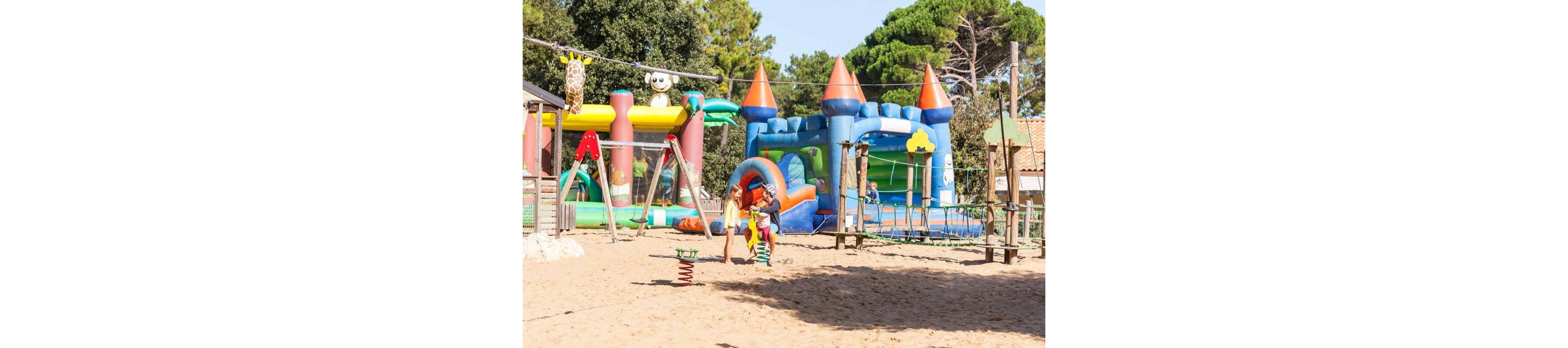 Two children walk together across a sandy outdoor playground filled with large inflatable structures. Brightly colored bouncy castles and slides with cartoon animal designs dominate the background. The scene is set on a sunny day with trees and small buildings surrounding the play area. The overall atmosphere is cheerful and playful, emphasizing fun and recreation for kids.