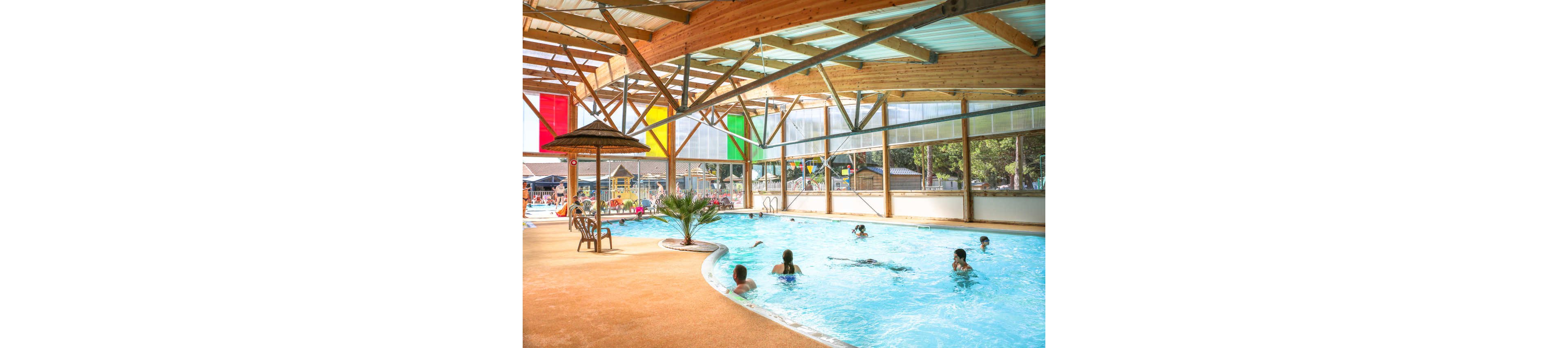 An indoor swimming pool is filled with people enjoying the water. Children and adults play and swim under a high wooden-beam roof with translucent panels. Large colorful vertical panels in red, yellow, and green hang along the side, letting in natural light. A small palm tree and thatched umbrella add a relaxed, vacation-like atmosphere to the poolside area.
