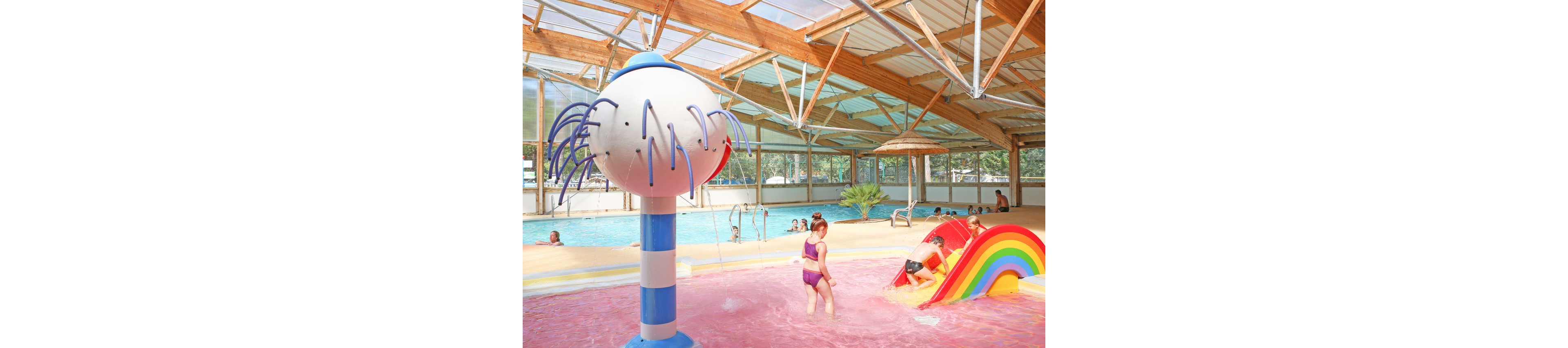 Young children play in a brightly colored indoor water park area. A girl walks through shallow pink-tinted water near a large white and blue water feature. Another child climbs a small rainbow slide that leads into the splash pool. In the background, adults and children relax in a larger swimming pool under a high wooden roof with large windows. The scene feels cheerful and family-friendly with playful aquatic structures and warm lighting.