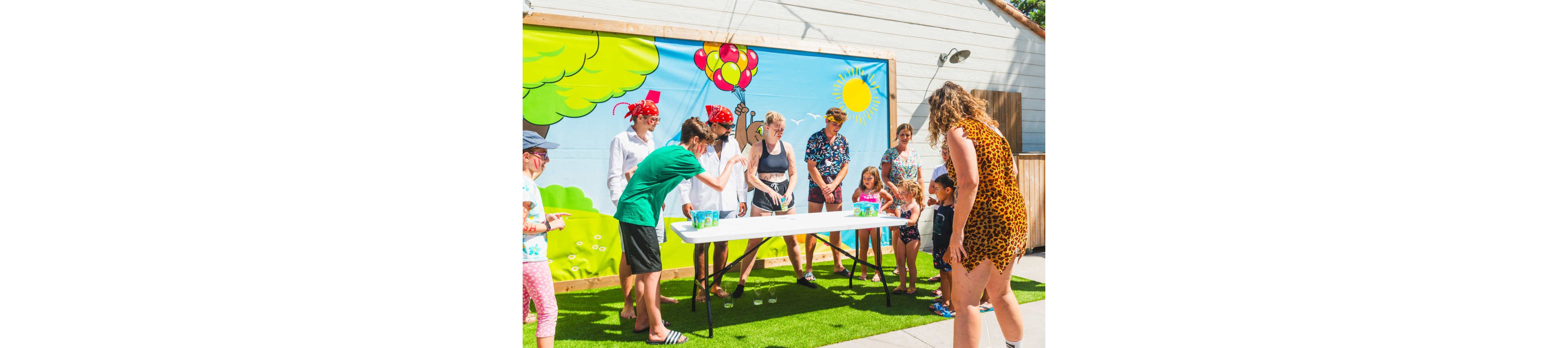 A group of adults and children gather around a white table at an outdoor party. The scene is set against a colorful cartoon mural featuring trees, balloons, and a bright sun. Kids watch as adults appear to organize a cup game on the table. Everyone is dressed in casual summer clothing, suggesting a warm, sunny day. The atmosphere feels playful and festive in a backyard or patio setting.