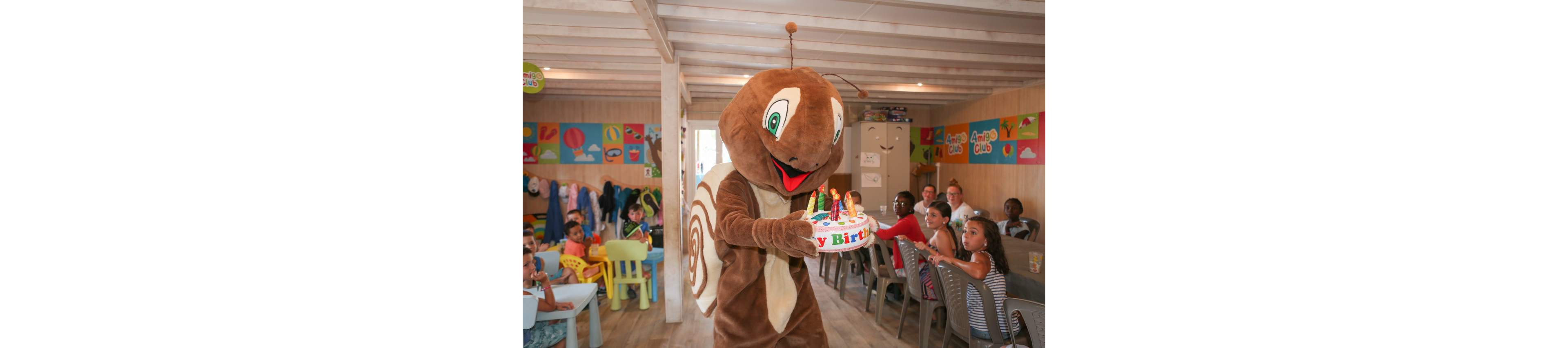 A person in a large brown snail mascot costume carries a birthday cake with colorful candles and decorations. The scene takes place in a bright indoor kids club or party room with wooden floors and walls. Children sit at tables and watch the mascot while adults supervise in the background. Colorful wall art and party decorations add to the festive atmosphere.