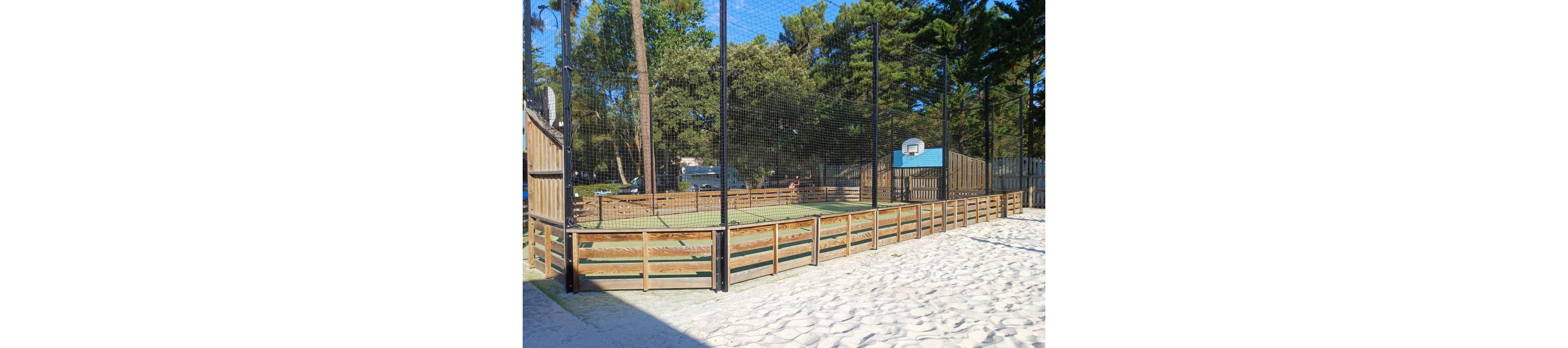 An outdoor multi-use sports court is enclosed by a wooden barrier and tall netting. The court surface appears to be a hard or artificial turf area suitable for ball games. A basketball hoop is mounted on the far fence, indicating the space can be used for basketball or similar activities. Surrounding the court are tall trees and wooden fencing, giving the scene a natural, recreational park atmosphere. In the foreground, a sandy area leads up to the edge of the court.