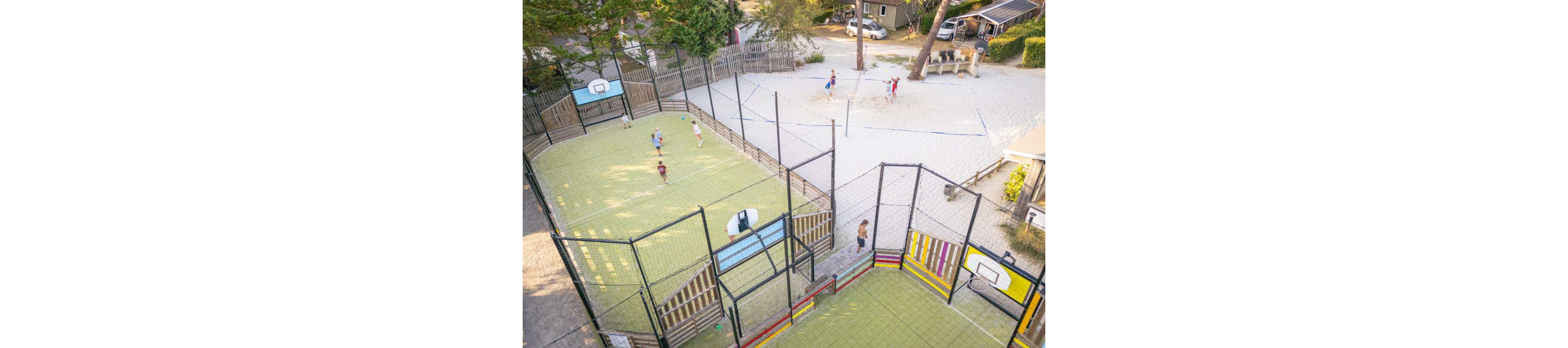 An aerial view shows children playing on a fenced multi-sport court next to a sandy volleyball area. The scene appears to be part of a recreational complex or holiday park surrounded by trees and small buildings. Kids are engaged in different games, including basketball and soccer, on the artificial turf court. The bright colors of the court walls and the warm daylight create a lively, energetic atmosphere.
