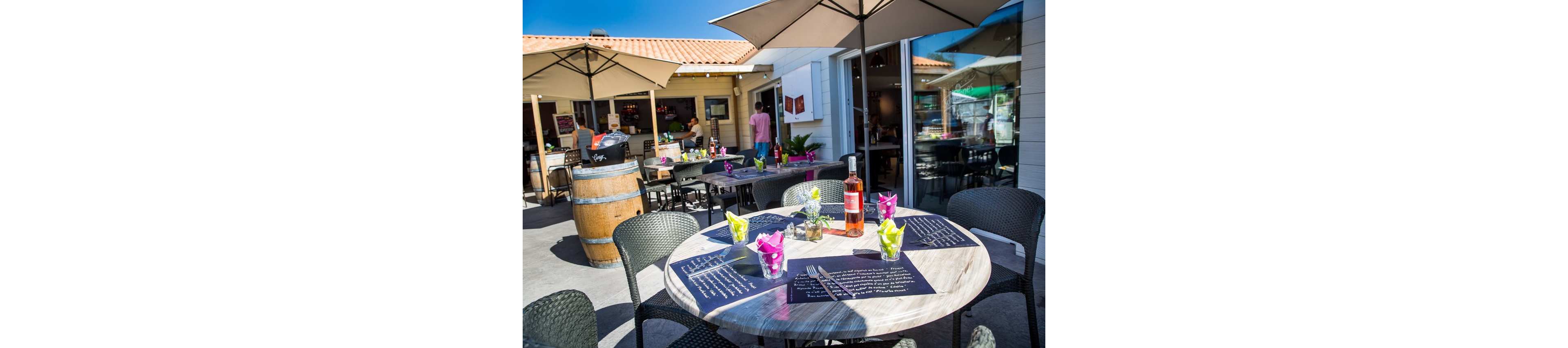 A round table is set at an outdoor café patio under large umbrellas on a bright sunny day. The table features a bottle of rosé wine, menus, cutlery, and colorful napkins in glasses. Other tables and wicker chairs fill the terrace, with a wine barrel and bar area visible in the background. People move around the open-air restaurant, creating a relaxed, social atmosphere.
