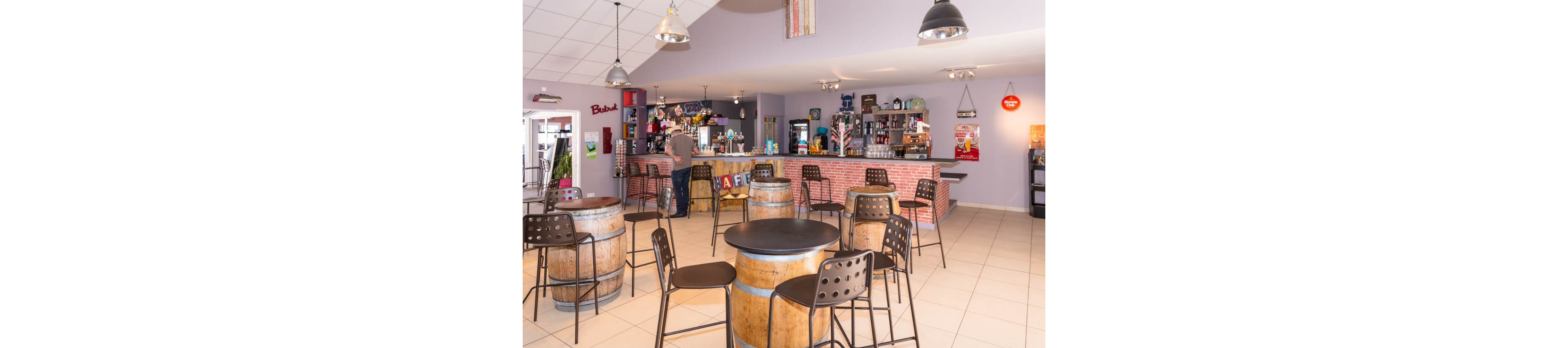 A bright bar interior features wooden barrels used as tables surrounded by metal chairs. A person stands at the long brick bar counter, which is lined with bottles, glasses, and colorful decorations. Pendant lights hang from the ceiling, illuminating the tiled floor and pastel-colored walls. The space feels casual and welcoming, suitable for social gatherings or relaxed drinks.