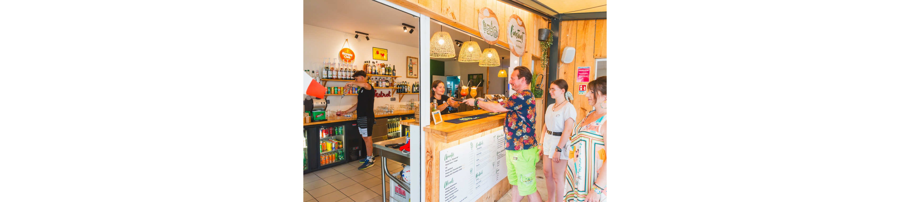 A group of people stands at a wooden bar counter receiving colorful drinks from a bartender. The bar interior features warm wooden panels, hanging wicker lamps, and shelves stocked with bottles and beverages. A staff member works behind a refrigerator filled with soft drinks, while a menu board is visible on the counter. The scene suggests a relaxed, casual atmosphere in a coastal or vacation setting.