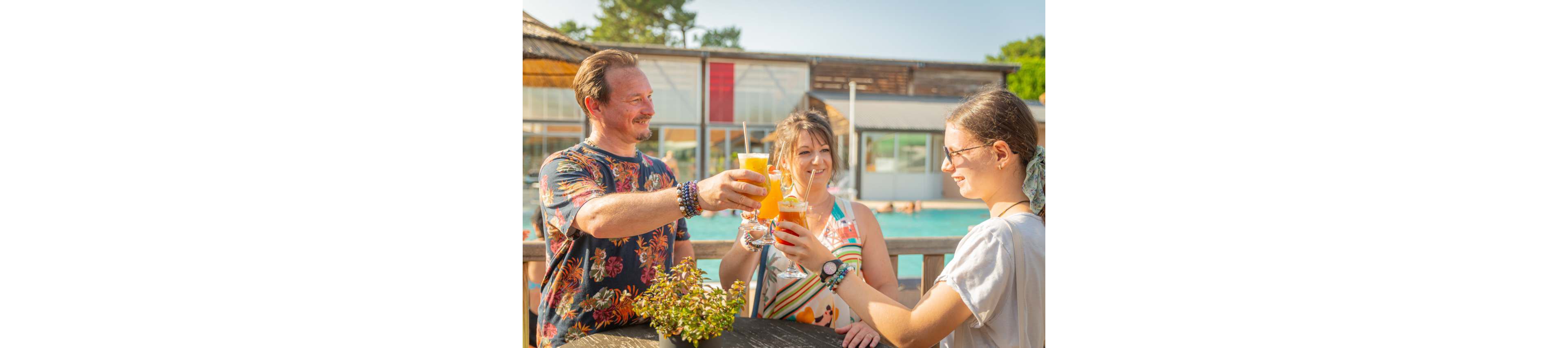 Three friends raise their drinks together while standing by an outdoor pool. The scene takes place on a sunny day at a leisure or resort area with wooden structures and clear blue water in the background. Each person holds a different bright beverage in tall glasses, suggesting refreshing summer cocktails or juices. Casual clothing, bracelets, and relaxed body language emphasize a cheerful vacation atmosphere.