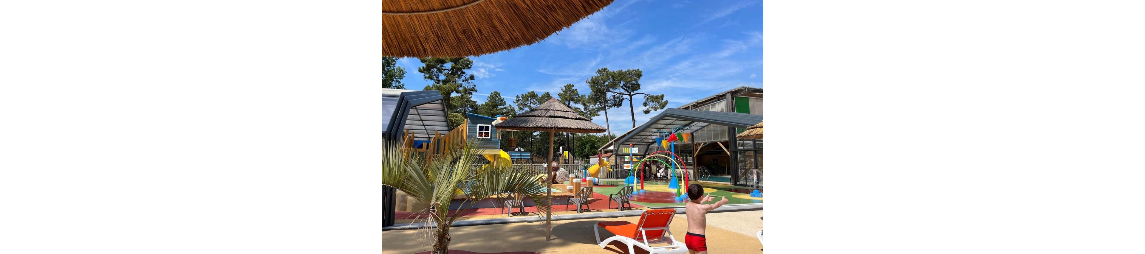 A colorful outdoor splash park features water play structures and slides under a bright blue sky. A young child in red swim trunks walks toward fountains and rainbow-shaped sprayers. Sun loungers and thatched umbrellas provide shaded seating around the play area. Palm plants and tall trees in the background create a relaxed, vacation-like atmosphere.
