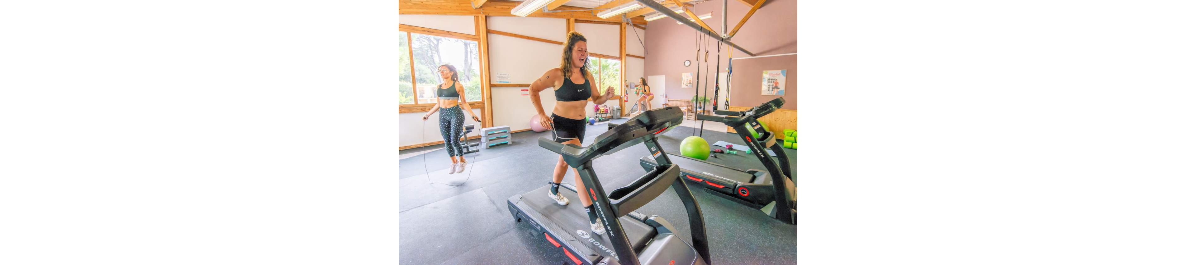 A woman runs on a treadmill in a bright gym while another woman jumps rope nearby. The fitness studio features large windows, wooden beams, and various exercise equipment including step platforms, stability balls, and resistance bands. Natural light fills the room, creating an energetic and motivating atmosphere. The scene emphasizes indoor cardio training and an active, healthy lifestyle.