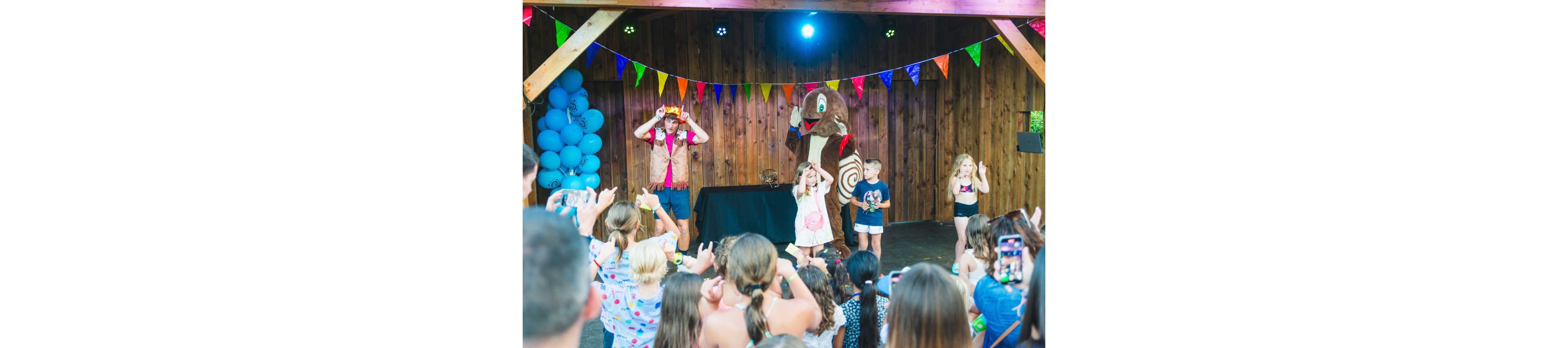 A group of children gathers in front of a small stage at an outdoor party or event. A performer in colorful clothes and a large costumed mascot entertain the kids, who are dancing and copying their gestures. Bright balloons, multicolored bunting, and stage lights decorate the wooden backdrop, creating a festive atmosphere. Parents and other attendees in the foreground hold up phones to capture photos and videos of the performance.