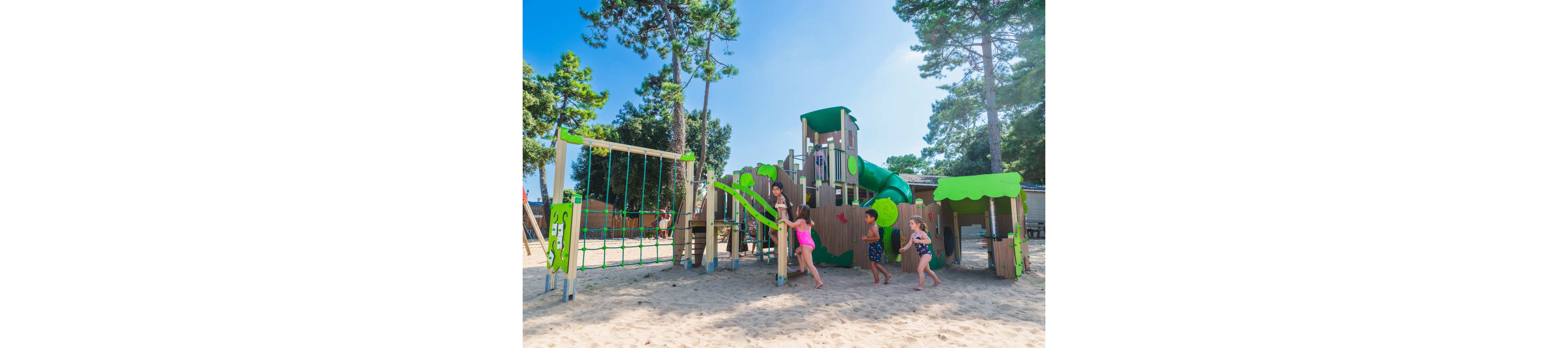 Several children play energetically on a large outdoor playground structure in a sandy area. The playground features slides, climbing frames, and rope elements in green and beige tones. Tall trees surround the play area, providing a natural backdrop under a bright blue sky. The scene feels lively and summery, with kids running, climbing, and exploring the equipment.