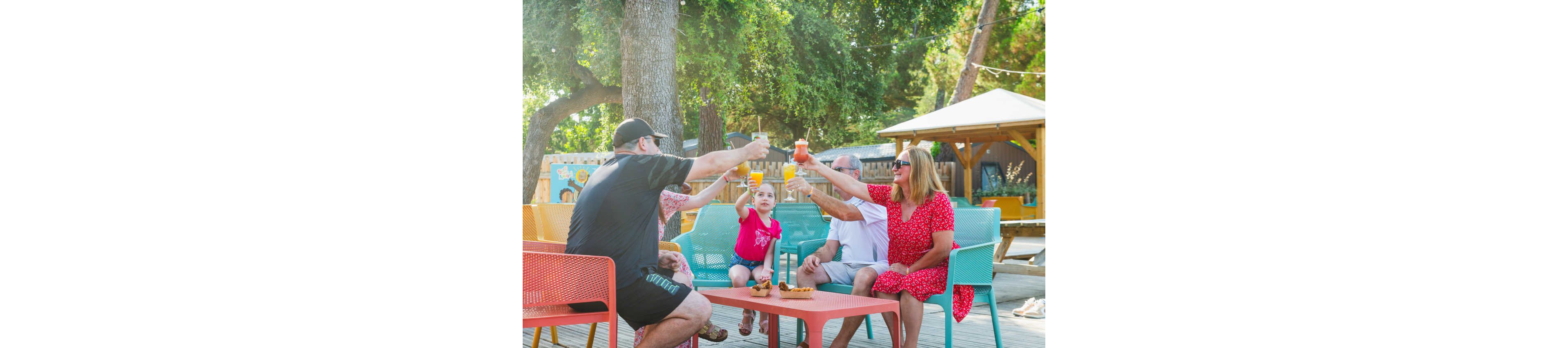 A family sits together at colorful outdoor tables, raising their drinks in a celebratory toast. The scene takes place in a sunny patio area shaded by large trees and surrounded by wooden structures. Adults and children share beverages and snacks, creating a relaxed, social atmosphere. Bright chairs and casual summer clothing add to the cheerful, vacation-like setting.