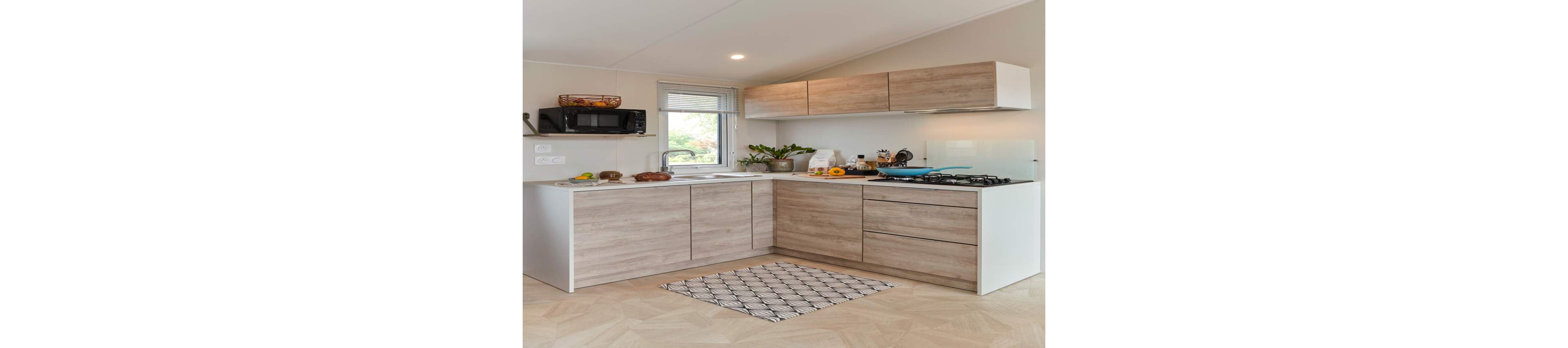 Contemporary L-shaped kitchen with light wood cabinets and white countertops is shown in a bright, compact space. A gas cooktop with blue pan, microwave, and sink are arranged along the walls beneath a window. Potted plants, cutting boards, and fresh produce add natural accents to the clean design. A patterned rug lies on the light herringbone floor, enhancing the cozy yet modern atmosphere.