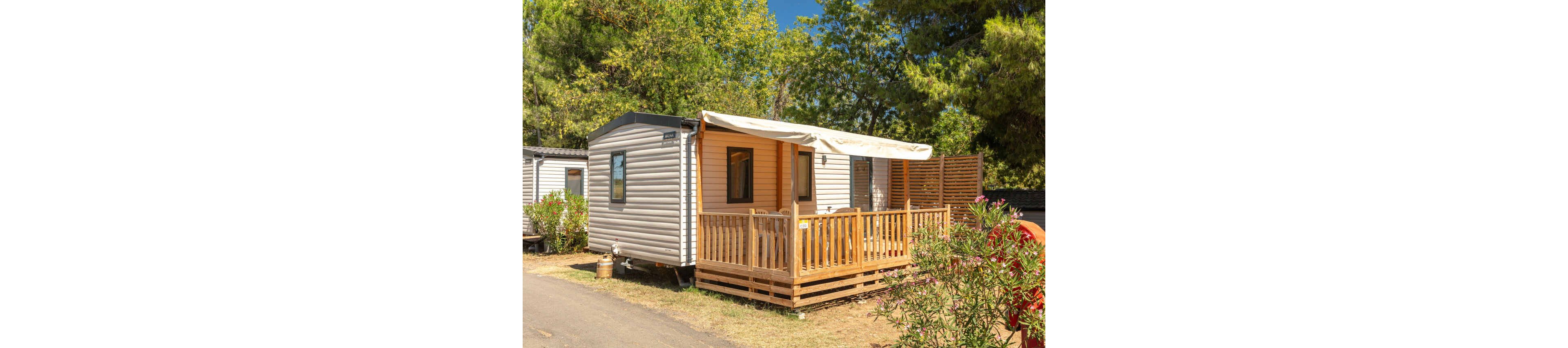 A compact mobile home with a wooden terrace is set in a sunny campsite. The structure features light-colored siding, a small covered porch, and large windows. Surrounding the home are green trees and shrubs, creating a relaxed outdoor atmosphere. The scene suggests a vacation setting with comfortable outdoor living space.