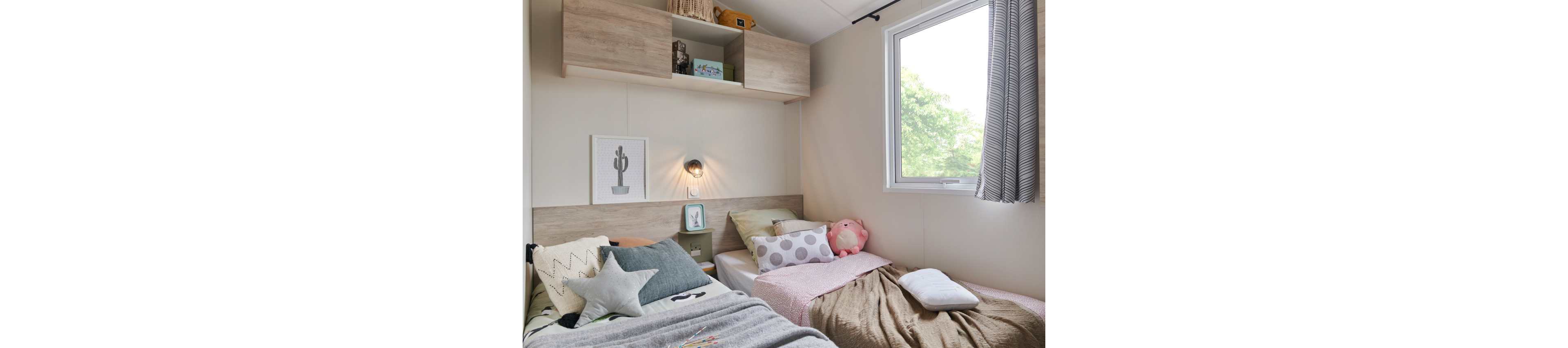 A small, cozy bedroom features two twin beds arranged in an L-shape, each with soft blankets and decorative pillows. The room is brightened by a large window with curtains, allowing natural light and greenery from outside to show through. Wall-mounted shelves hold books and decor, while a framed cactus print and a bedside lamp add personality. Plush toys and patterned cushions suggest a child-friendly, comfortable space designed for rest and play.