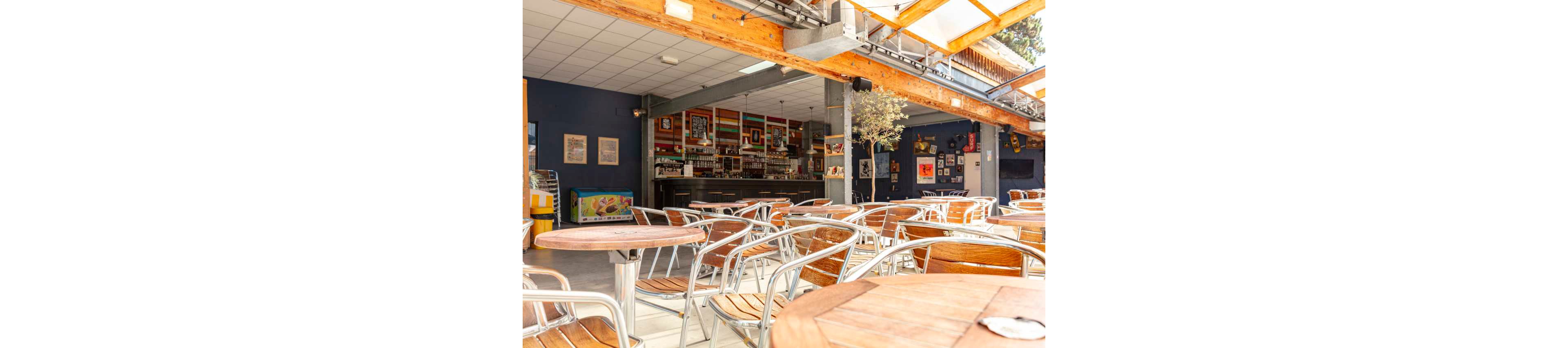 An empty café terrace features rows of round wooden tables with metal chairs under a partially covered roof. Warm sunlight streams in from the open side, creating bright highlights and soft shadows across the seating area. In the background, a bar counter is lined with bottles and decorated with framed posters and artwork on dark blue walls. The overall atmosphere feels relaxed and inviting, suggesting a quiet moment before customers arrive.