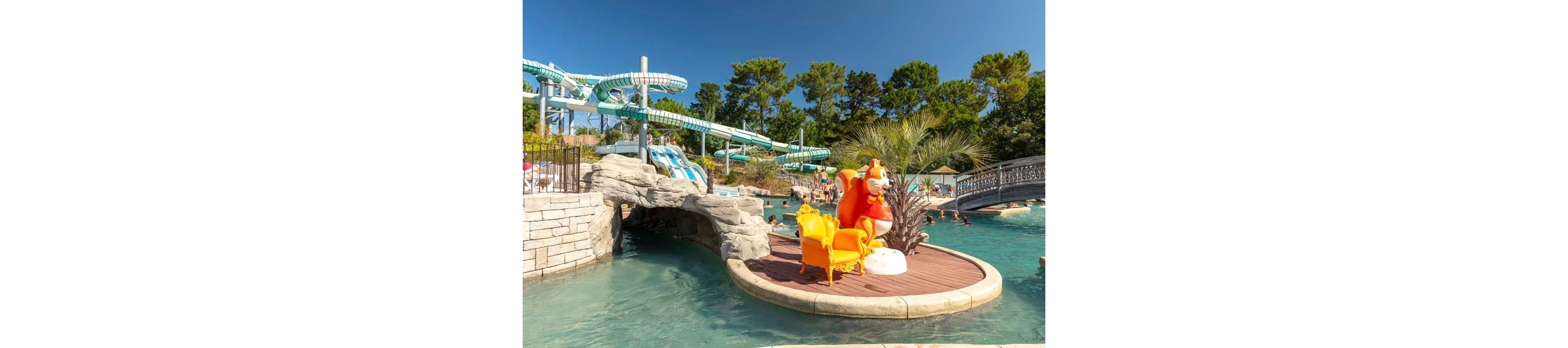 An outdoor water park features winding blue and white water slides surrounded by tall green trees. In the foreground, a small island deck holds bright yellow chairs and an orange cartoon mascot figure next to a palm tree. Swimmers enjoy the clear pool water near a rocky tunnel and a decorative bridge. The sunny weather and vibrant colors create a fun, family-friendly atmosphere.