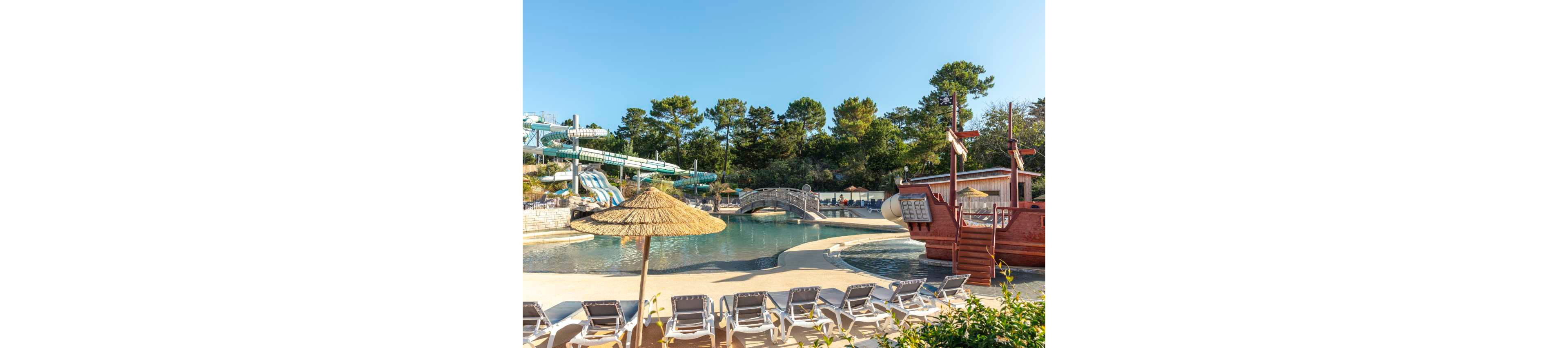 An outdoor water park features a shallow lagoon-style pool with a red pirate ship play structure. Empty sun loungers line the poolside under straw parasols, suggesting a relaxing holiday setting. Curving water slides and a small arched bridge add to the playful atmosphere. Tall green trees surround the area, creating a natural backdrop under a clear blue sky.