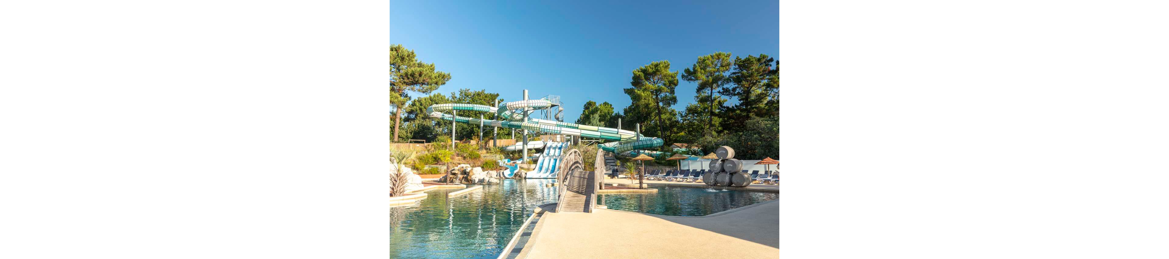 An outdoor water park features multiple large twisting water slides emptying into a clear blue pool. A small bridge crosses part of the pool, leading toward lounge chairs and umbrellas arranged along the deck. Tall green trees surround the area, creating a natural backdrop under a bright blue sky. The scene suggests a sunny, relaxing day at a family-friendly aquatic attraction.