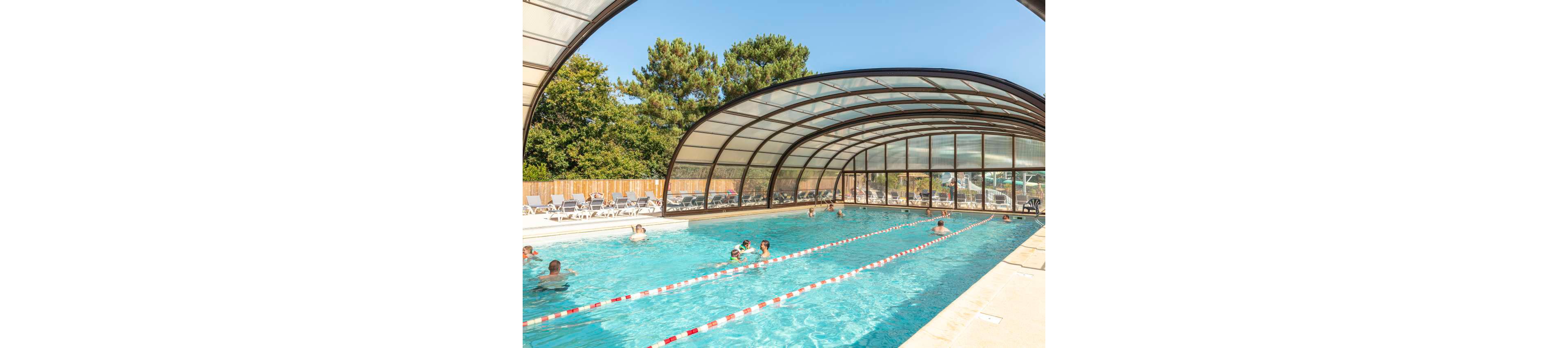 A large covered swimming pool features several people enjoying the water. The transparent arched roof lets in sunlight while providing shelter over the pool. Lane dividers stretch the length of the pool, and lounge chairs line the deck along a wooden fence. Trees and greenery surround the area, creating a relaxed vacation atmosphere under a clear blue sky.