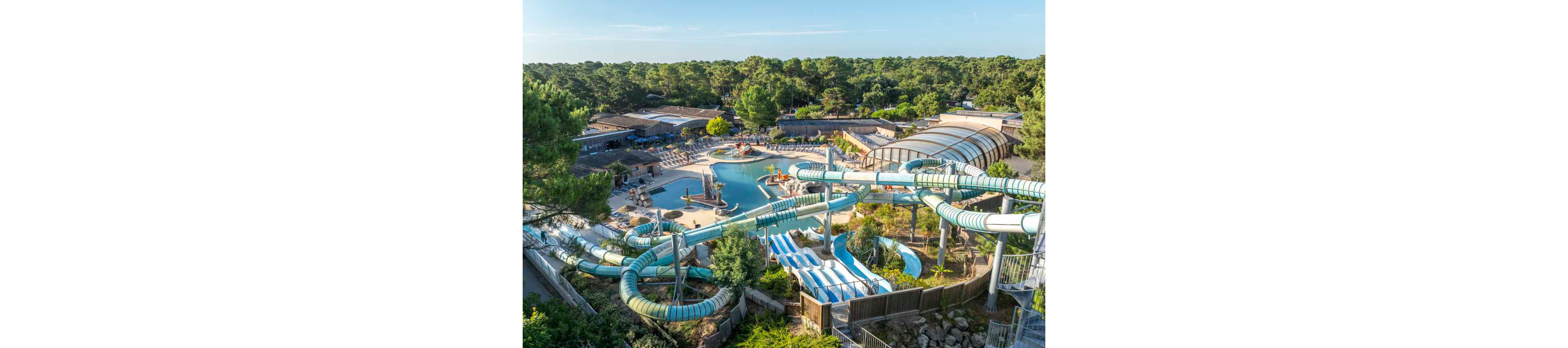 A large outdoor water park is shown from above, featuring multiple twisting water slides and a central swimming pool. The complex includes sun loungers, shaded seating areas, and a partially covered pool structure. Surrounding the park is a dense forest of green trees, creating a secluded holiday atmosphere. The scene suggests a sunny day at a family-friendly resort with various aquatic attractions.