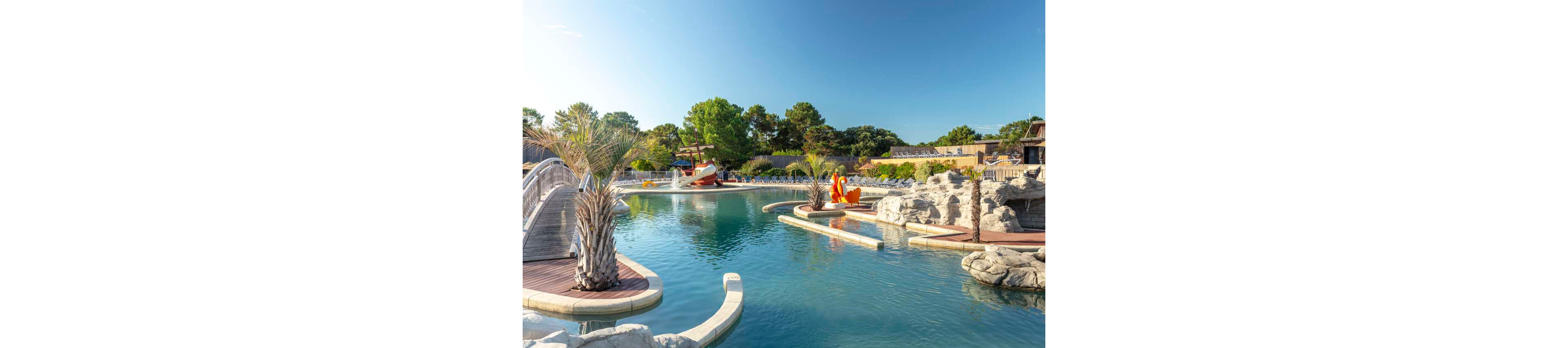 An outdoor resort-style pool features clear blue water surrounded by rocks and palm trees. A small pirate ship play structure and colorful statues create a playful, family-friendly atmosphere. Wooden bridges and curved walkways cross over the water, adding visual interest and pathways for visitors. The scene is lit by bright sunshine under a clear blue sky, suggesting a warm vacation destination.