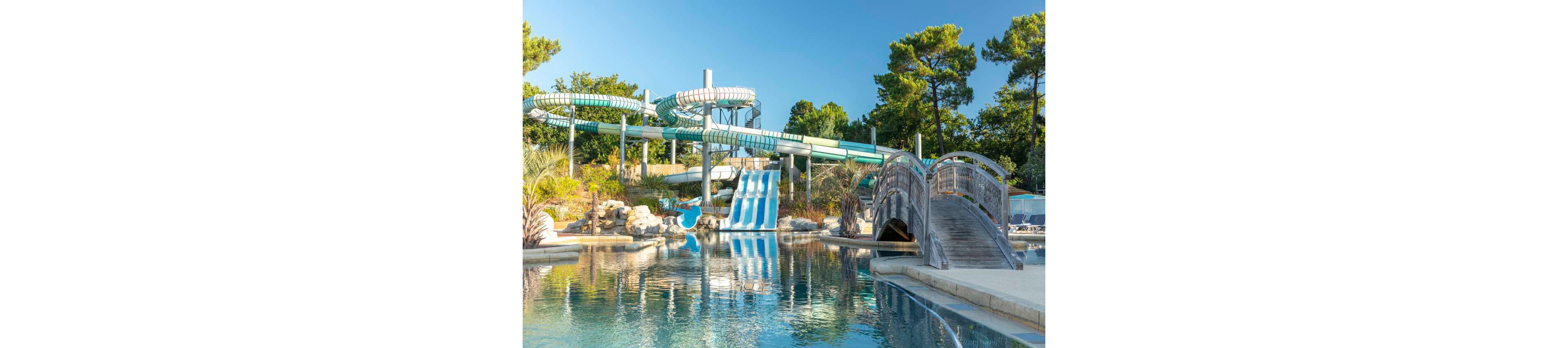 An outdoor water park features multiple large twisting water slides and a set of blue and white racing slides. A wooden footbridge arches over a calm pool in the foreground, reflecting the surrounding structures. Lush greenery, rocks, and tall trees frame the aquatic area under a clear blue sky. The scene suggests a relaxing yet adventurous summer recreation setting.