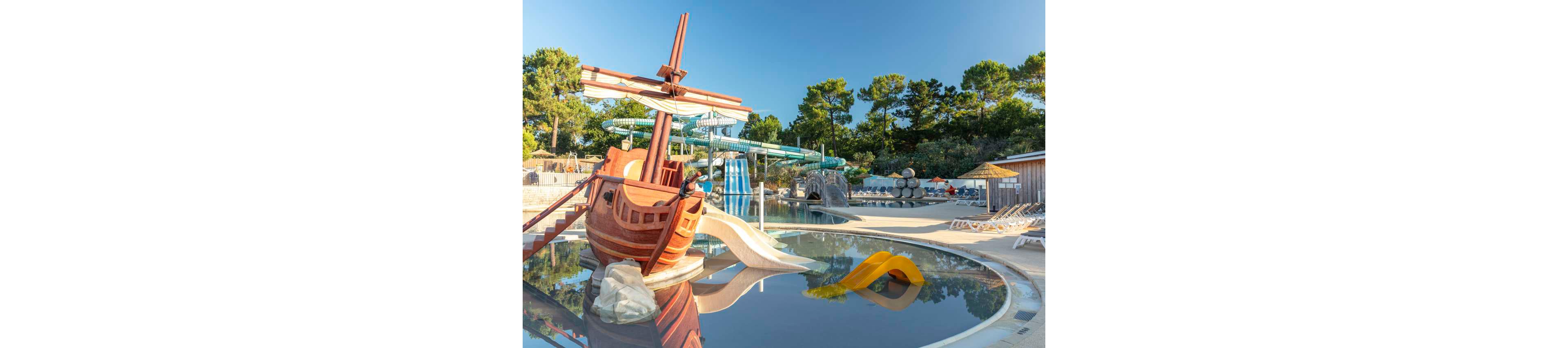 An outdoor water park features a large wooden pirate ship play structure beside a shallow pool. Multiple colorful water slides and tube slides curve in the background, set among tall green trees. Empty lounge chairs line the poolside under clear blue skies, suggesting a calm, off-peak moment. The scene evokes a family-friendly, summer holiday atmosphere focused on fun and relaxation.