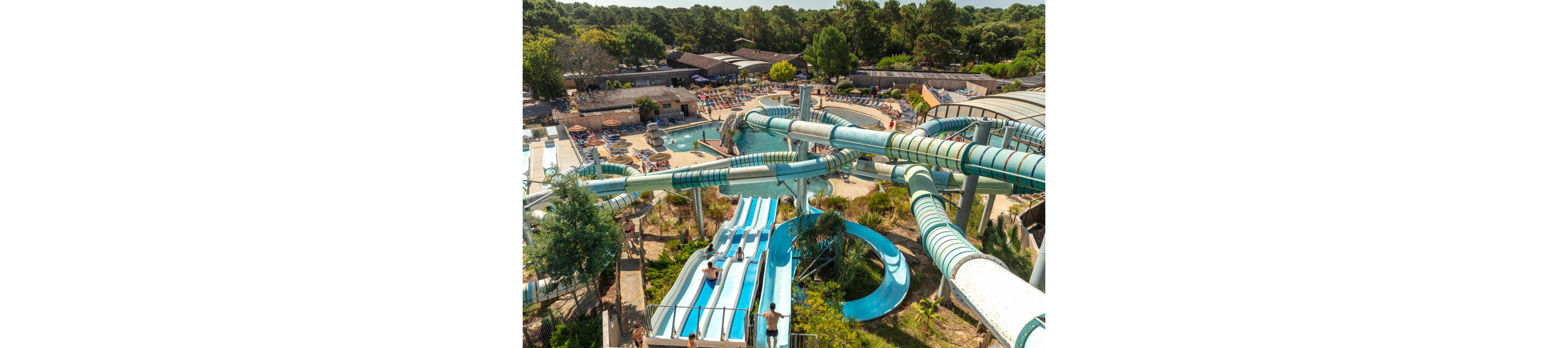 An aerial view shows a large outdoor water park with multiple twisting water slides and pools. People are enjoying the sunny day, lounging on deck chairs and swimming in the central pool area. Several riders are captured mid-descent on open and enclosed slides surrounded by greenery. Buildings and dense trees frame the park, emphasizing a lively vacation atmosphere.