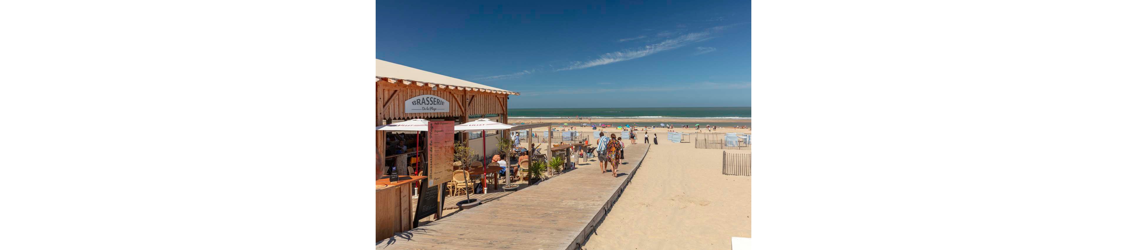 A wooden beachside brasserie sits beside a wide sandy boardwalk leading toward the ocean. People walk along the boardwalk in summer clothing while others relax at outdoor café tables under umbrellas. Colorful beach tents and sunbathers are scattered along the sand near the shoreline. The sky is clear and blue, creating a bright, relaxed seaside atmosphere.