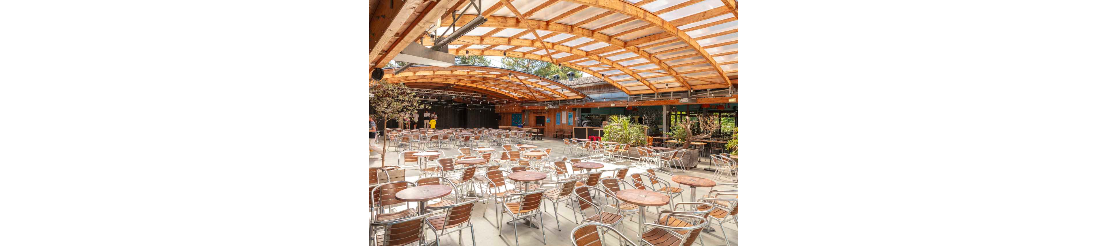 An expansive open-air café terrace is filled with rows of empty metal and wood tables and chairs. A large curved wooden canopy with transparent panels shelters the seating area while still letting in natural light. In the background, a few people stand near a bar or service counter surrounded by plants and colorful walls. The scene suggests a relaxed social venue prepared for guests, possibly for dining, drinks, or events.
