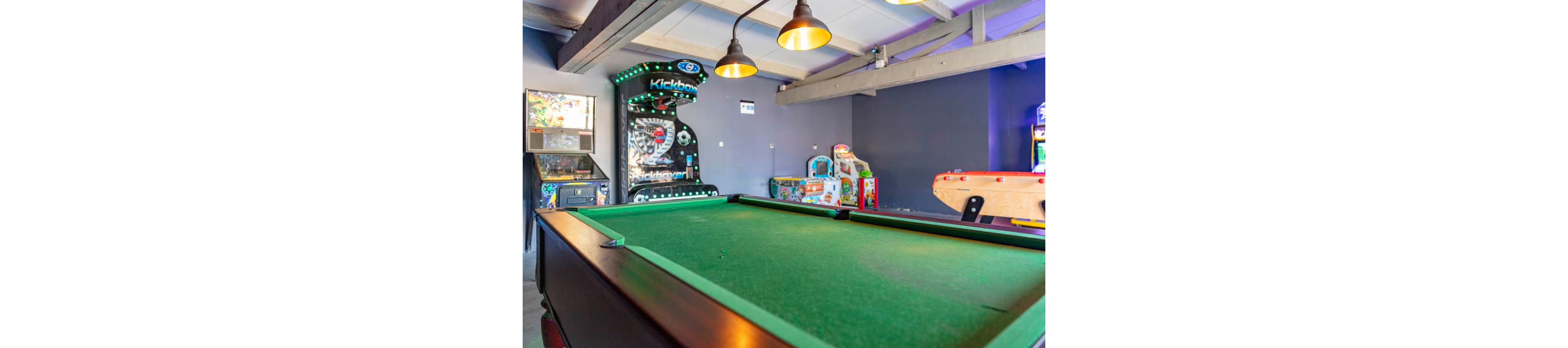 A green felt pool table dominates the foreground of a bright recreation room. In the background, several arcade machines and a foosball table line the walls, creating a fun entertainment space. Overhead industrial-style pendant lights hang from exposed beams, casting warm light across the room. The overall scene suggests a casual, playful environment for games and socializing.